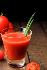 A glass cup with tomato juice, next to it lies a branch of tomatoes. On a wooden table there is a glass of tomato juice and tomatoes lie on a black background.