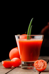 A glass cup with tomato juice, next to it lies a branch of tomatoes. On a wooden table there is a glass of tomato juice and tomatoes lie on a black background.
