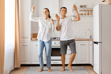 Full length portrait of man and woman having fun together in the kitchen, young adult couple is dancing at home, raised arms, smiling, expressing happiness.