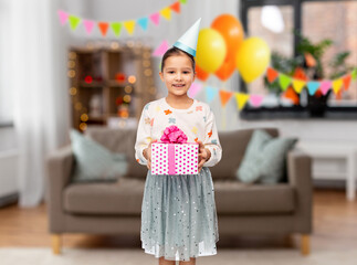 birthday, childhood and people concept - portrait of smiling little girl in party hat with gift box decorated living room background