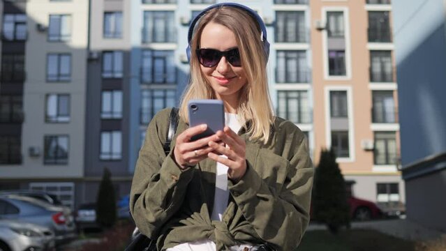 Beautiful blond smiling girl wearing casual clothes and sunglasses choosing music from app playlist. Young woman listening to music on her smartphone in wireless headphones enjoy good quality sound.