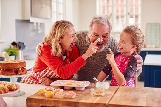 Grandparents With Granddaughter Baking Cupcakes And Putting Icing On Their Noses At Home