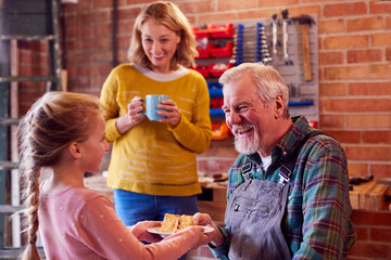 Granddaughter With Grandmother Bringing Grandfather Cake And Hot Drink In His Home Workshop