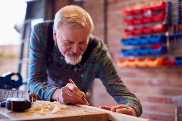 Senior Male Carpenter In Garage Workshop Measuring And Marking Piece Of Wood With Pencil