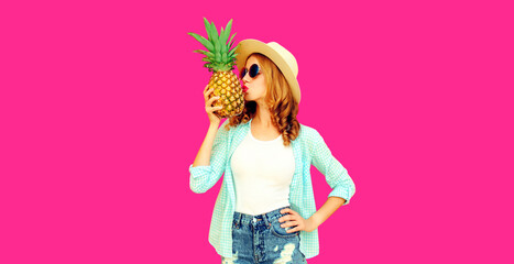 Summer portrait of stylish woman kissing tropical pineapple wearing straw hat, sunglasses on pink background