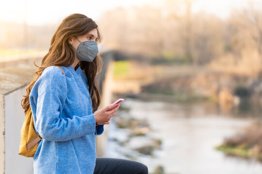 Young Girl With Gray Face Mask Looking At The Phone While Walking In The Park