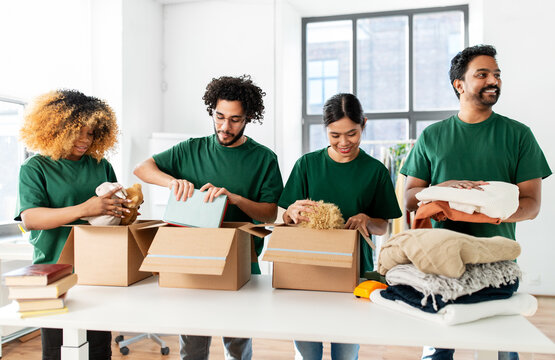 charity, donation and volunteering concept - international group of happy smiling volunteers packing clothes and other stuff in boxes at distribution or refugee assistance center