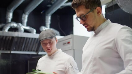 Chef checking ingredients in restaurant kitchen and talking to male colleague as he taking notes during food inventory