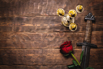 Ancient knight sword, burning candle and red rose flower on the wooden flat lay table background with copy space.
