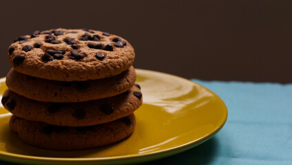 Pile of chocolate chip cookies on a yellow plate and on a blue tablecloth and dark background
