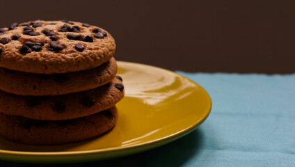 Pile of chocolate chip cookies on a yellow plate and on a blue tablecloth and dark background