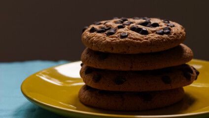 Pile of chocolate chip cookies on a yellow plate and on a blue tablecloth and dark background
