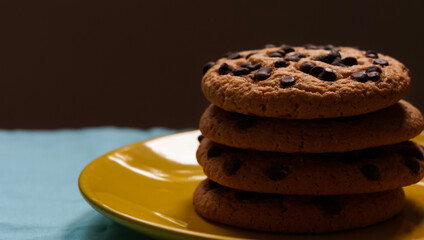 Pile of chocolate chip cookies on a yellow plate and on a blue tablecloth and dark background