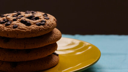 Pile of chocolate chip cookies on a yellow plate and on a blue tablecloth and dark background