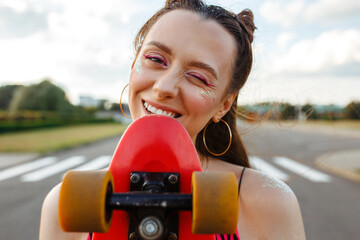 Optimistic young teen girl skater with glistening makeup biting skateboard and standing on road in summer city while looking at camera   © Dzmitry