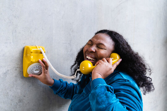 Woman talking through old-fashioned telephone receiver laughing in front of gray wall