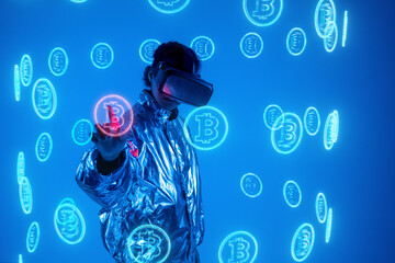 Woman wearing virtual reality simulator touching bitcoin symbol on screen against blue background