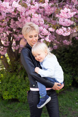 Fototapeta premium Young mother mom holding her little baby son boy child under blossoming Sakura Cherry trees with falling pink petals and beautiful flowers