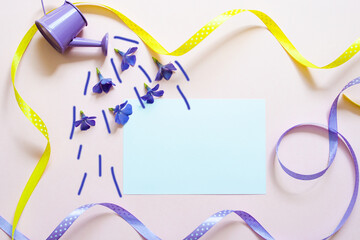 White sheet on a pink background with lilac and yellow ribbons. A watering can pours a shower of periwinkle flowers onto a leaf. Romantic message concept with copy space