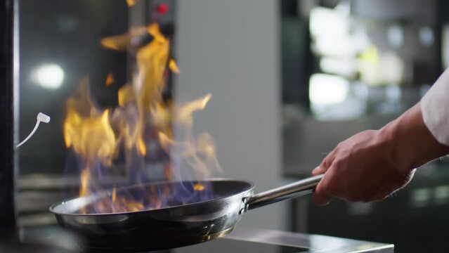 Slow motion close up shot of unrecognizable restaurant chef using lighter to start fire in skillet and flambeing vegetables while cooking in kitchen