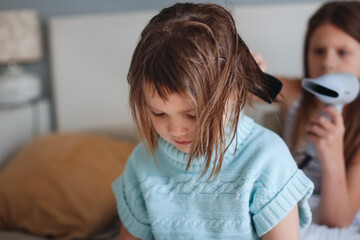 Fototapeta premium Little sister blow-drying her little sister's hair. Children in the bedroom on the bed, siblings take care of each other