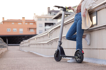 Cropped view of unrecognizable young woman leaning on the railing holding a electric scooter town scape view behind