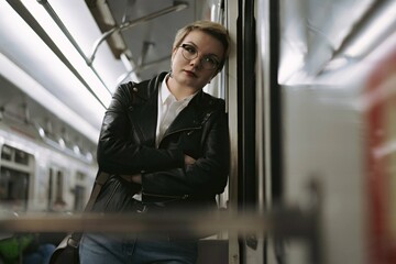 young caucasian short haired blond woman wearing leather jacket, jeans and glasses leaned on door in car of metro, looking aside. Image with selective focus