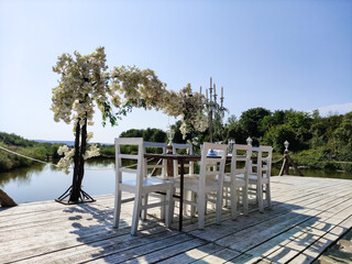 Fototapeta na wymiar Decorative arch with white flowers.Wedding decoration on the shore of a lake.Table with white chairs placed next to the archway with white flowers