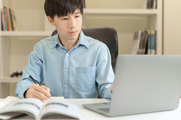 Young Asian college male student writing notes, using laptop for learning online education, business learning, watching online webinar. Focused man doing homework. Online education learning concept.