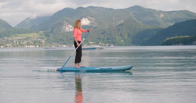 Woman paddles on sup board at Wolfgang mountain lake, Austria, early morning. Active tourism and travel, water sport during summer holidays.