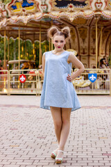 A beautiful girl in a blue dress stands against the background of a carousel