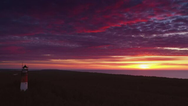 Beautiful sunset over green forest. Slow drone flight. View of the lighthouse, zoom out. Sea in the distance. Orange sun, pastel pink clouds, red sky.