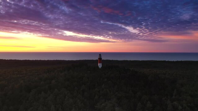 Beautiful sunset over green forest. View of the lighthouse. Orange sun, pastel pink clouds. Sea in the distance.