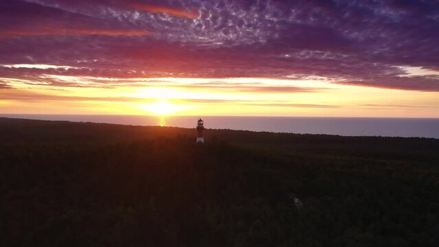 Beautiful sunset over green forest. View of the lighthouse. Orange sun, pastel pink clouds, red sky. Slow drone flight.