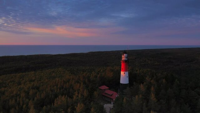 Aerial view of the lighthouse during sunset. Sea in the background. Blue sky and orange clouds. Slow drone flight over the green forest.
