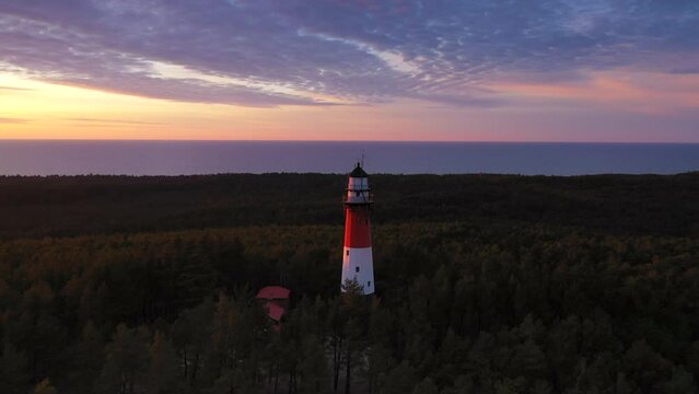 Sunset over green forest. Sea in the background. View of the lighthouse, zoom out. Orange sun, pastel pink clouds. 