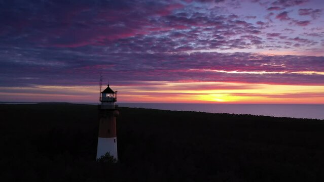 Beautiful sunset over green forest. View of the lighthouse, sea in the background. Orange sun, pastel pink clouds, red sky.
