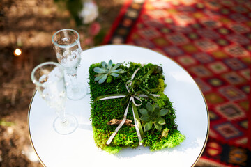 Wine glasses, wedding rings bedding of grass and moss on white table next to bohemian tipi arch...