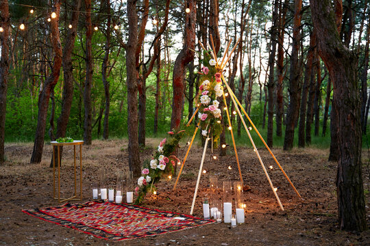 Bohemian tipi arch made of wooden rods decorated with pink roses, candles on carpet, pampass grass, wrapped in fairy lights on outdoor wedding ceremony venue in pine forest. Floristic compositions.