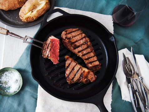 Two Grass Fed Steaks in a Cast Iron Skillet with Baked Potatoes in the Background.