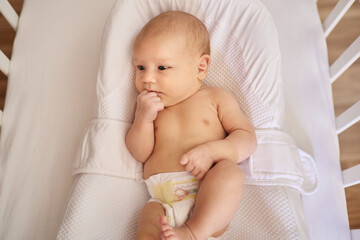 a newborn baby lies in a cocoonababy in a diaper in a white bed with slatted walls lies and pulls the fist into his mouth