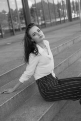 A young brunette girl in white shirt and striped trouser is sitting on the stairs