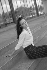 A young brunette girl in white shirt and striped trouser is sitting on the stairs