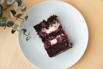 Piece of chocolate sponge cake with sweet cherry and cream cheese filling on white plate. Flat lay. Wooden background
