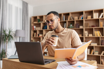 Middle eastern male student learning remotely, reading book, browsing internet on laptop and drinking coffee at home