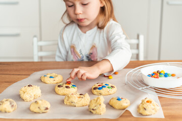 Little caucasian girl puts   multi-colored candies in shortbread cookies dough. Making cookies at home