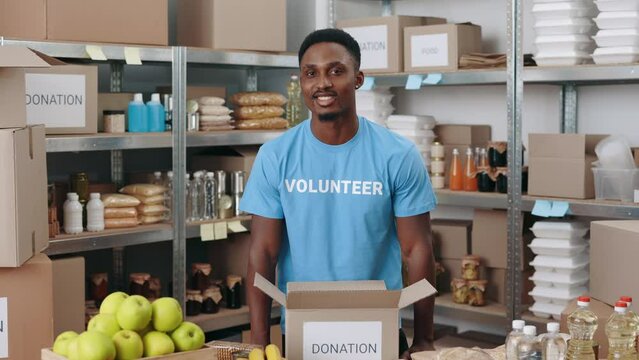 Handsome young guy in blue volunteer t-shirt preparing cardboard boxes for donation at warehouse. Portrait of african american volunteer posing indoors.