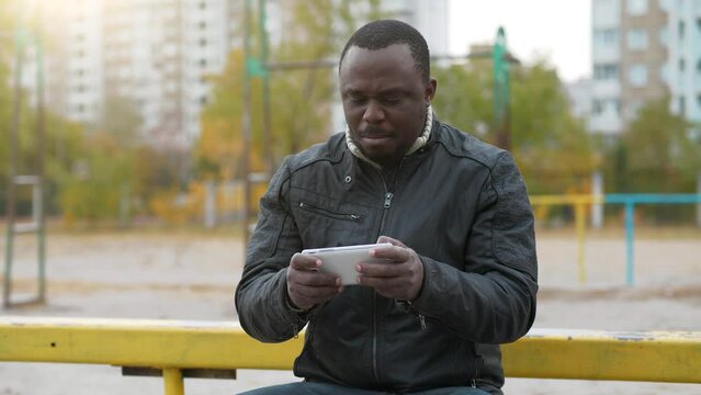African american man sits at sport ground and plays the game on his smartphone
