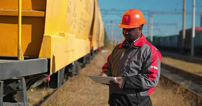 Inspector of wagons at freight train station makes notes in his tablet computer. African american railway man with tablet-pc at freight train terminal. Cinema 4K 60fps video