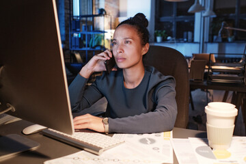 Concentrated young businesswoman in black shirt sitting at table with papers and talking by phone while discussing charts on computer at night